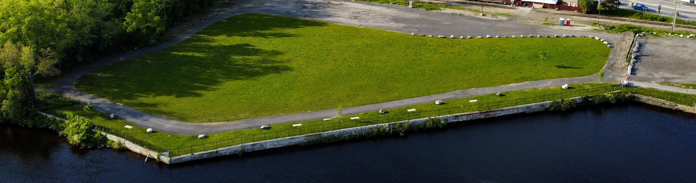 The Worumbo lawn from above, a wide green field edged by a path and the stone river wall, with the Androscoggin River in front